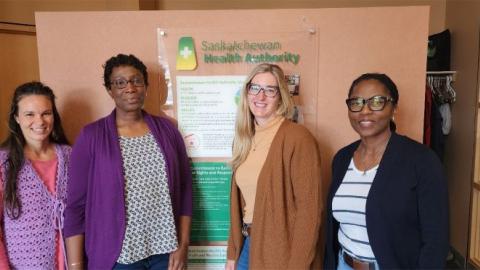 Four women standing side-by-side in front of a wall. On the wall, a bulletin identifying the Saskatchewan Health Authority supports for Mental Health and Addictions services, accessible through calling HealthLine 811.