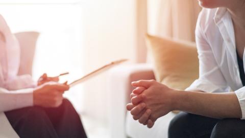 Two people in conversation, seated and facing each other. One person is holding a clipboard or writing tablet, while the other person sits across from them, leaning in with hands clasped.