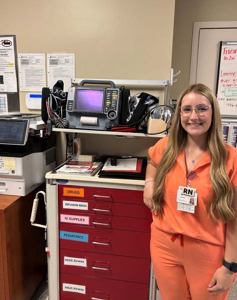 ICU nurse Ashlyn Dyok wears orange scrubs and smiles while standing next to a supply cart in a hospital room.