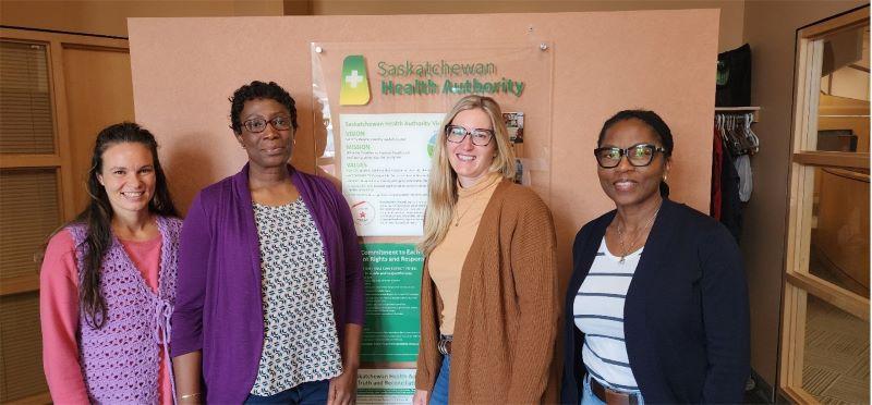 Four women standing side-by-side in front of a wall. On the wall, a bulletin identifying the Saskatchewan Health Authority supports for Mental Health and Addictions services, accessible through calling HealthLine 811.